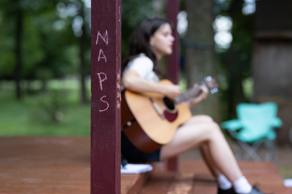 In the foreground, the word NAPS is written on a post. Blurred in the background is Azalea, playing the guitar.