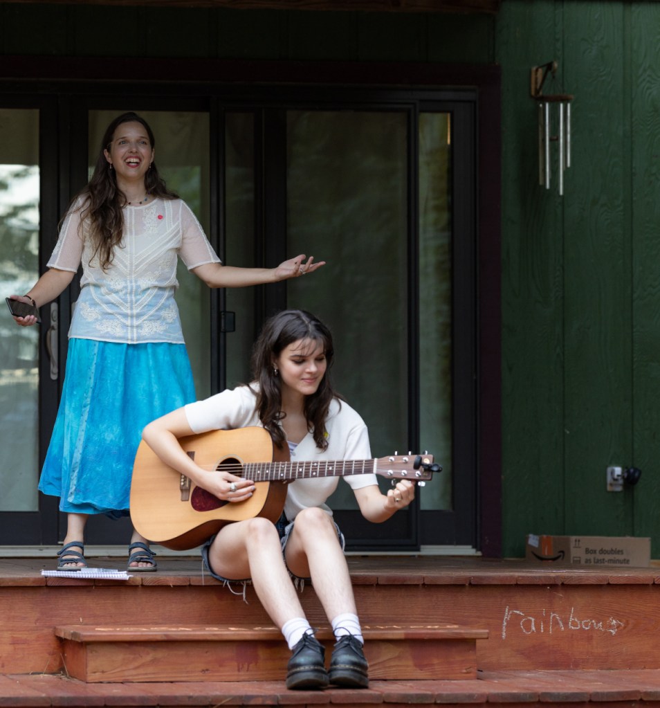Artistic director Aili Huber welcomes the crowd. In the foreground, musician Azalea Twining tunes her guitar.