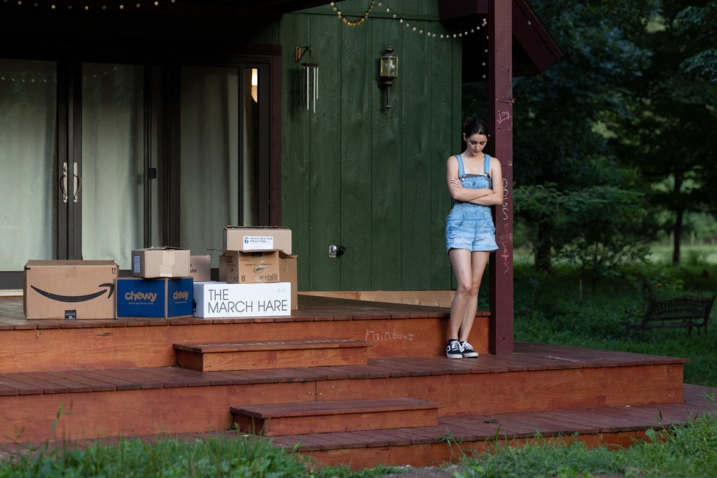 Katie stands alone on stage, leaning against a post. A pile of boxes is behind her.