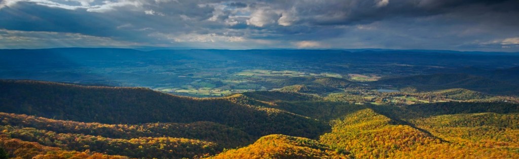 Panorama of the Blue Ridge Mountains, with orange fall trees in the foreground, blue mountains at the horizon, and sun slanting through gray clouds